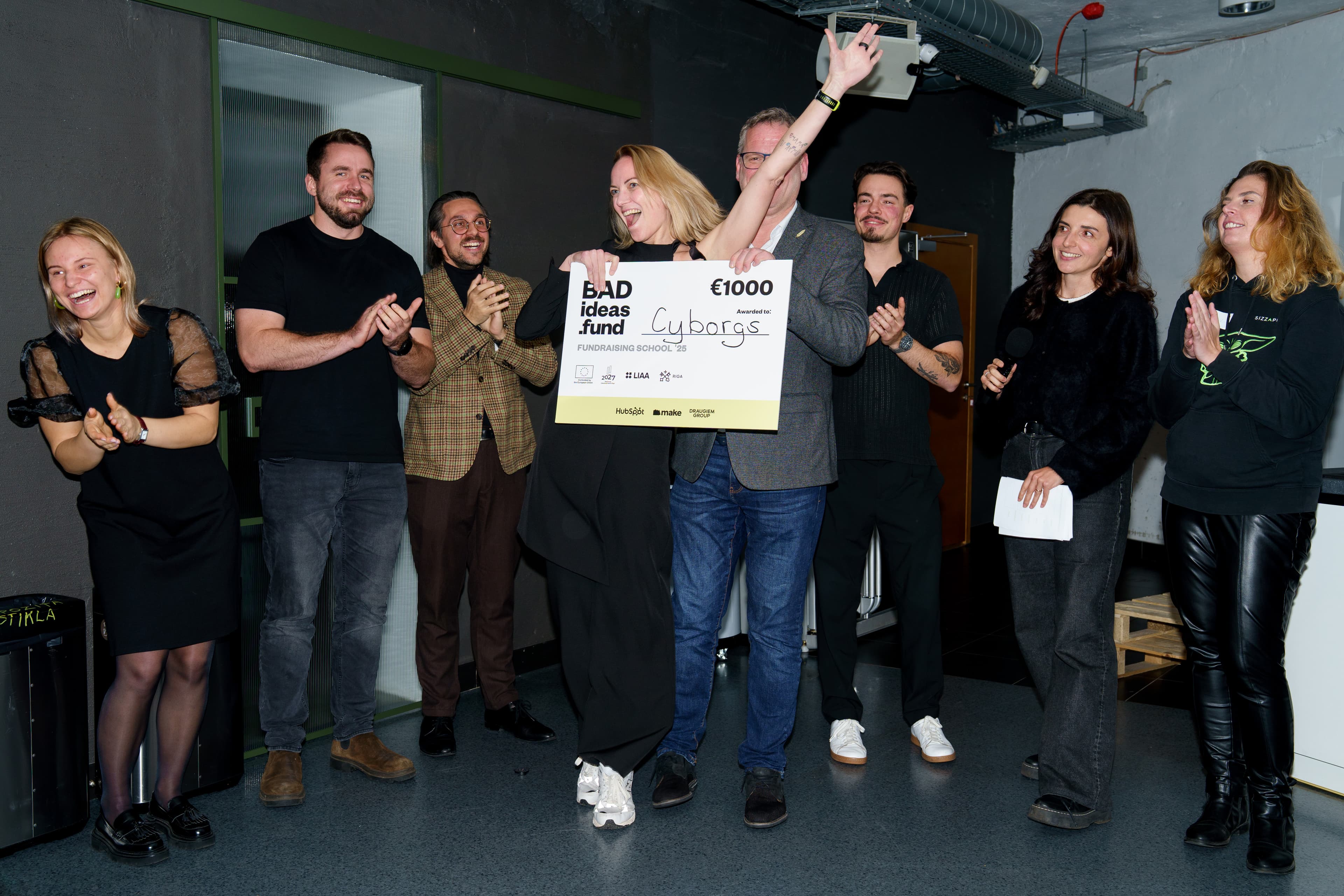Team Cyborgs holds up a €1,000 prize cheque at the BADideas.fund Fundraising School 2025 final while participants applaud.